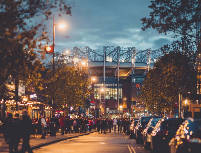 Crowds walking towards a lit stadium on event night with cars parked on nearby streets